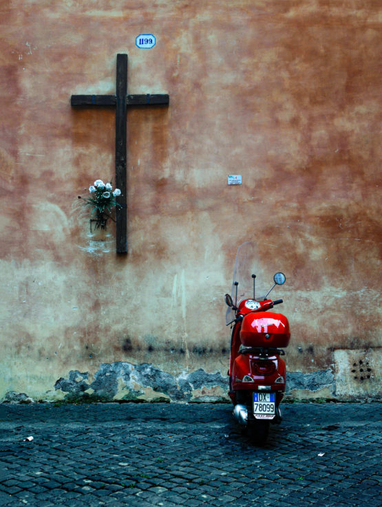 Een brommer staat onder een groot kruis op straat geparkeerd